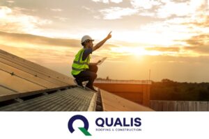Worker in safety gear crouches on rooftop solar panels, inspecting with a tablet and pointing towards the sky during sunset. "QUALIS Roofing & Construction" logo displayed at the bottom of the image. Choose QUALIS for energy-efficient roofing that truly matters.