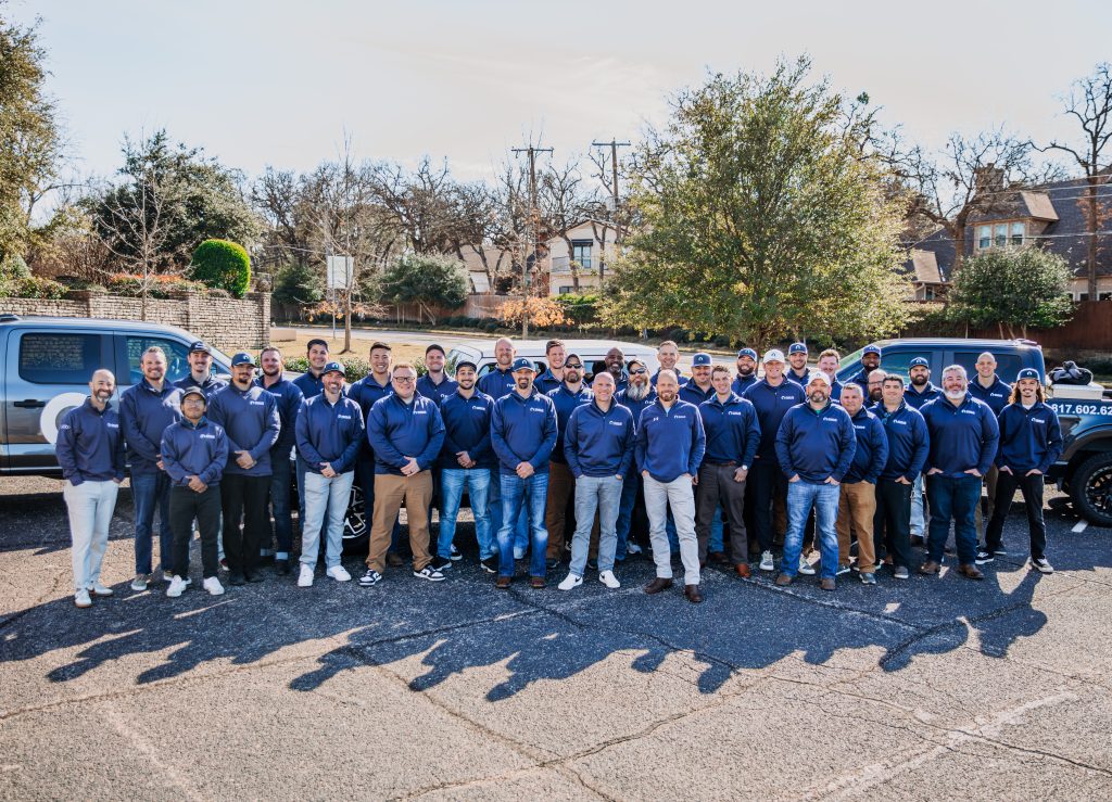 A group of about 40 people wearing matching blue jackets stands together outdoors in a parking lot, with two trucks and trees in the background.