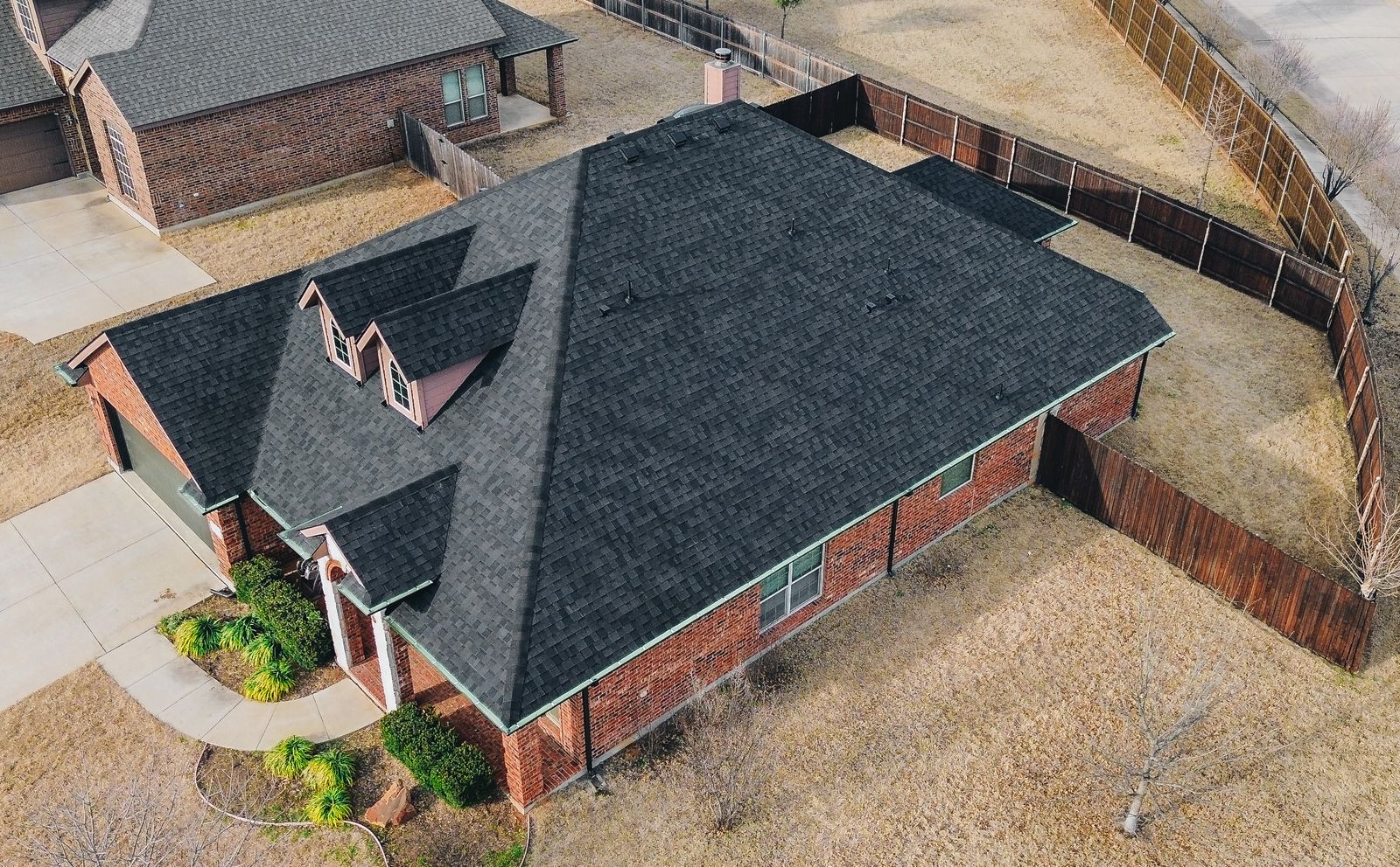 Aerial view of a single-story brick house with dark shingles installed by a leading Dallas roofing contractor, fenced backyard, driveway, and dry grass lawn in a suburban neighborhood.