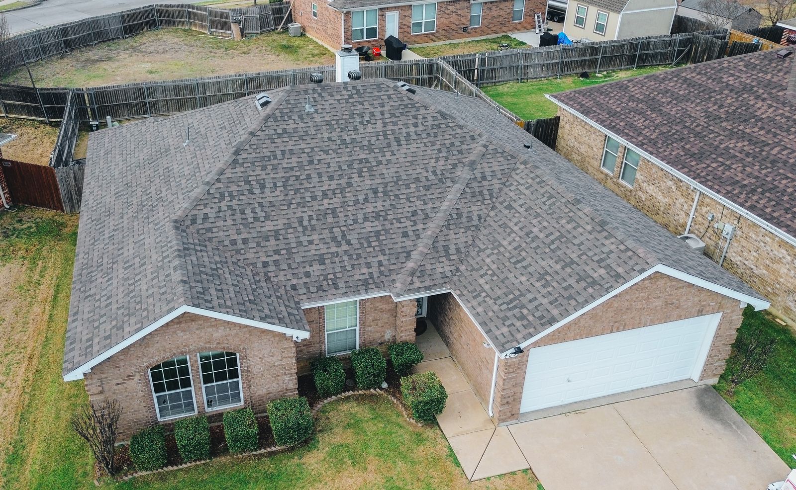 Aerial view of a single-story brick house with a gray shingle roof, white garage door, small front porch, and neatly trimmed bushes—showcasing quality residential roofing by a trusted Dallas roofing contractor.